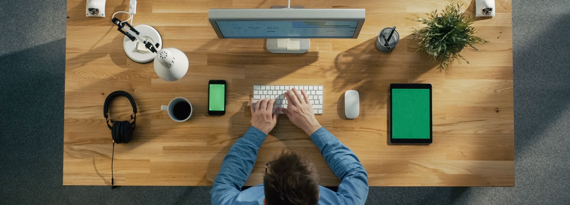 Een man zit aan zijn bureau achter zijn computer. Rechts van hem ligt een tablet en links van hem zijn mobiele telefoon en een kop koffie.